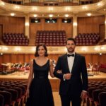 Pár v eleganckom oblečení vstupuje do koncertnej sály s červenými sedadlami a osvetlenou scénou.
